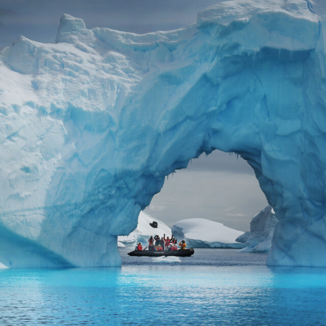 A small Zodiac inflatable boat carries tourists beneath a huge blue iceberg in the lagoons and bays surrounding the Antarctic peninsular